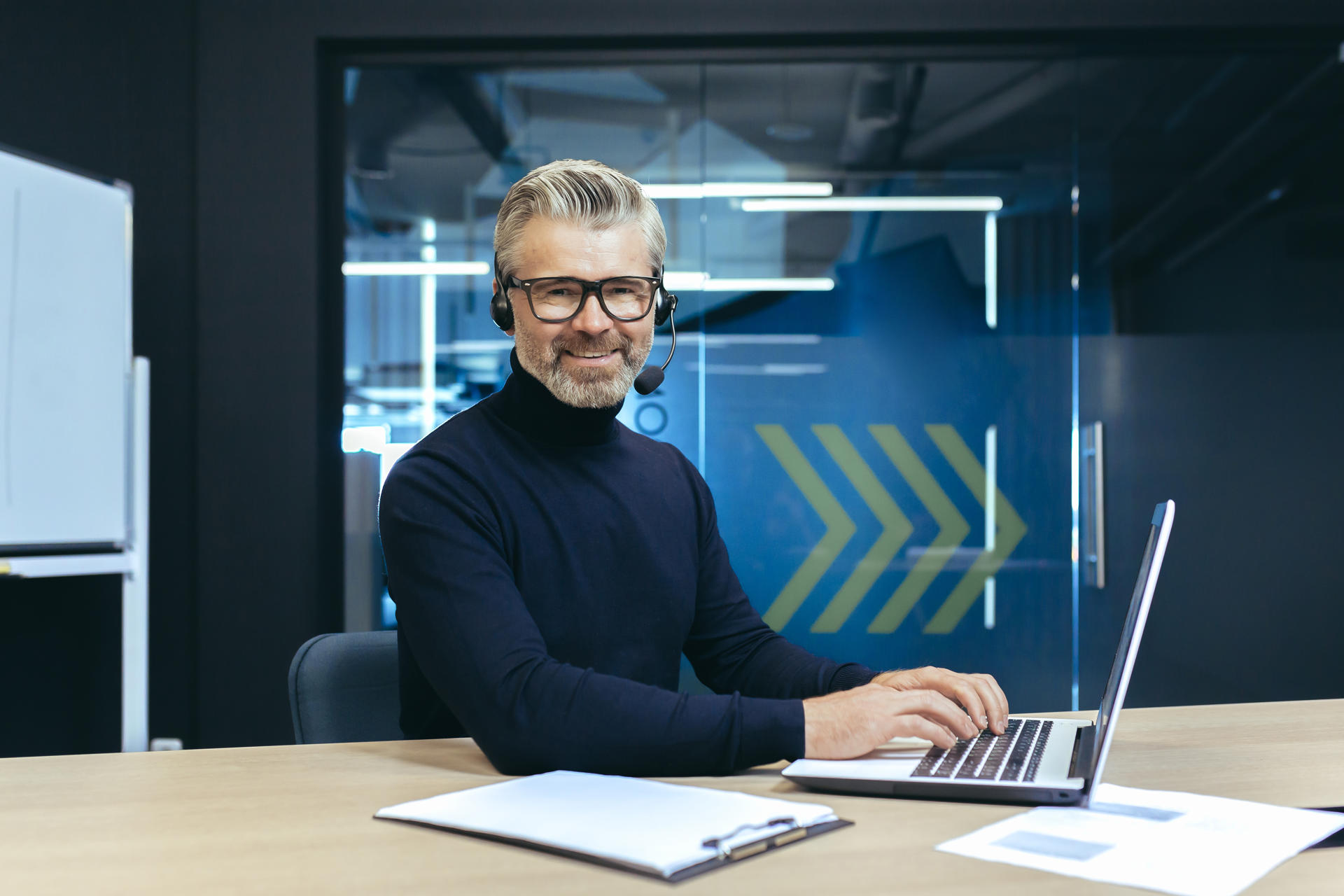 A gray-haired handsome businessman with a headset and a microphone is working on a laptop. Sits in the office at the table, conducts video conferences, consults. He looks at the camera, smiles.
